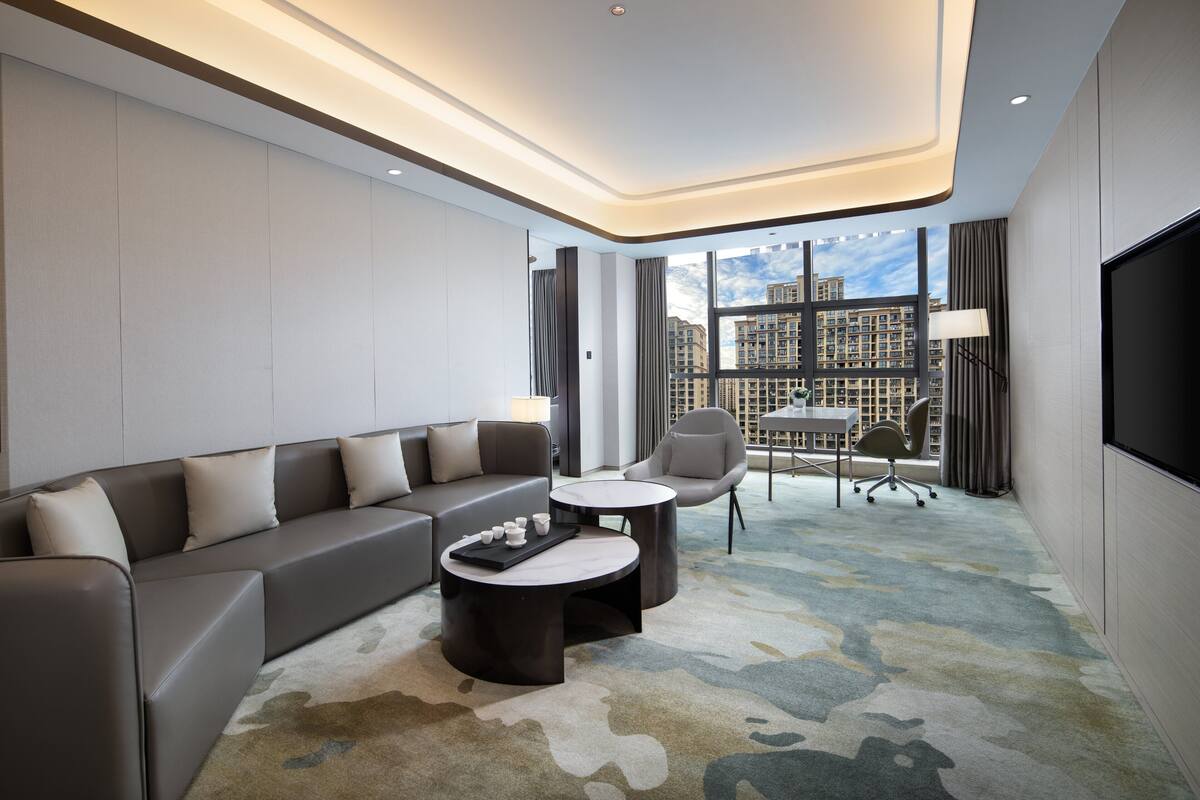 Ein modernes Hotelzimmer mit einem grauen Sofa, zwei Sesseln, einem Couchtisch und einem Fernseher.