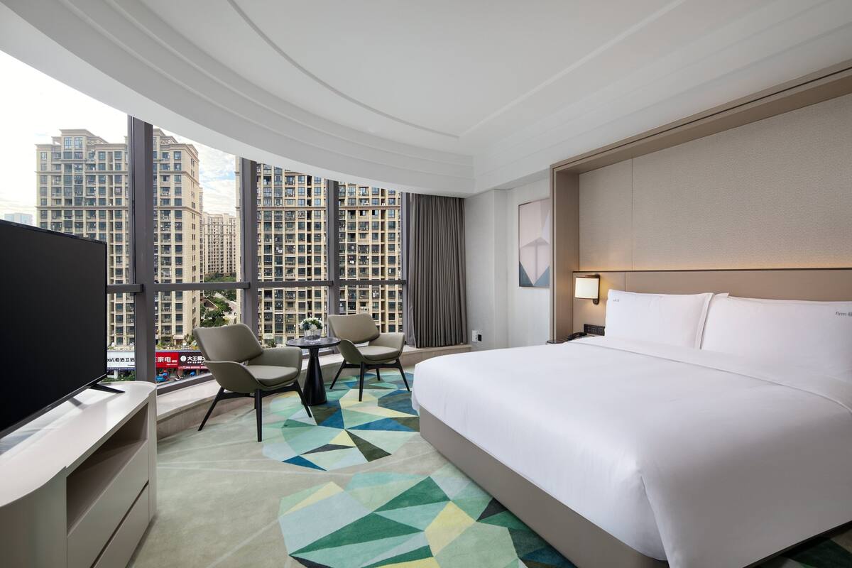 Ein Hotelzimmer mit einem großen Bett, einem Schreibtisch mit Flachbildfernseher, zwei Stühlen, einem kleinen Tisch mit einer Vase und Blick auf hohe Gebäude durch große Fenster.