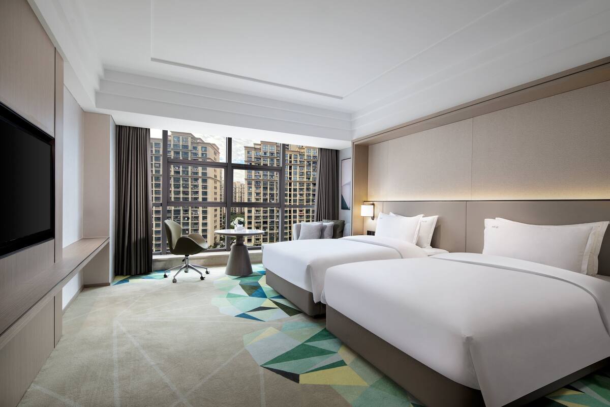 Ein Hotelzimmer mit zwei Betten, einem Fernseher, einem Schreibtisch und Blick auf hohe Gebäude.