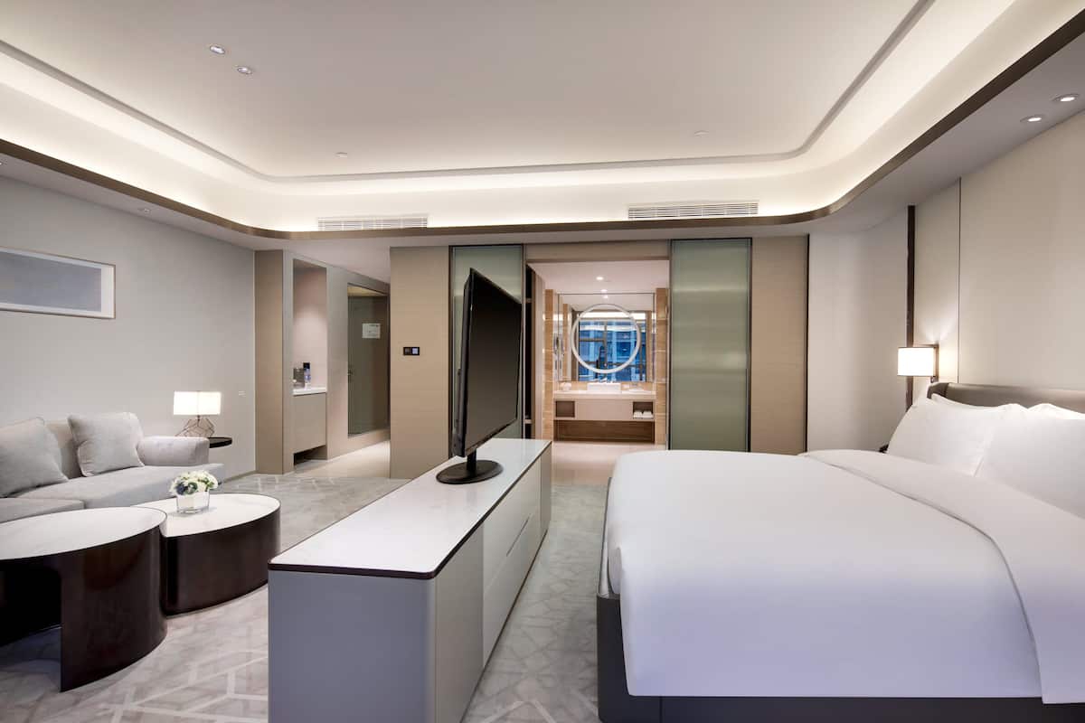 Ein modernes Hotelzimmer mit einem großen Bett, einem Schreibtisch, einer Couch und einem durchgehenden Türspalt erkennbaren Badezimmer.