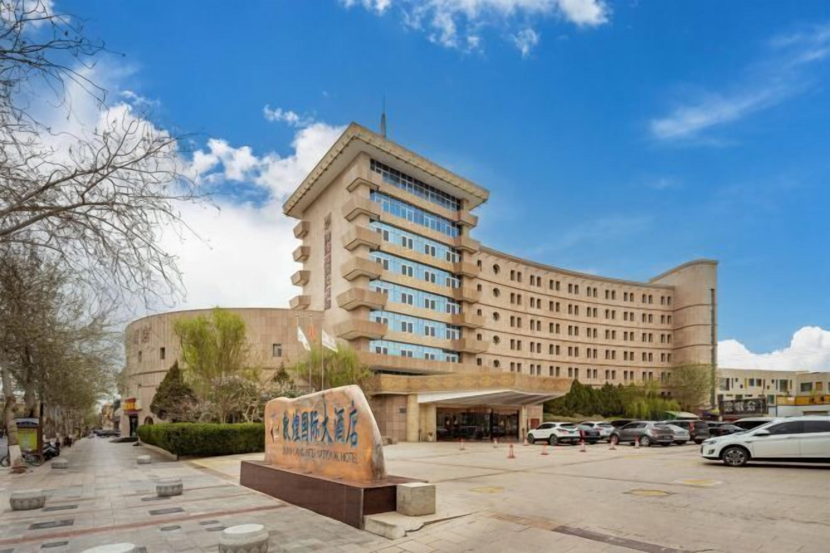 A large, multi-story hotel with a curved facade, a prominent entrance with a covered area, and a parking lot with several cars.