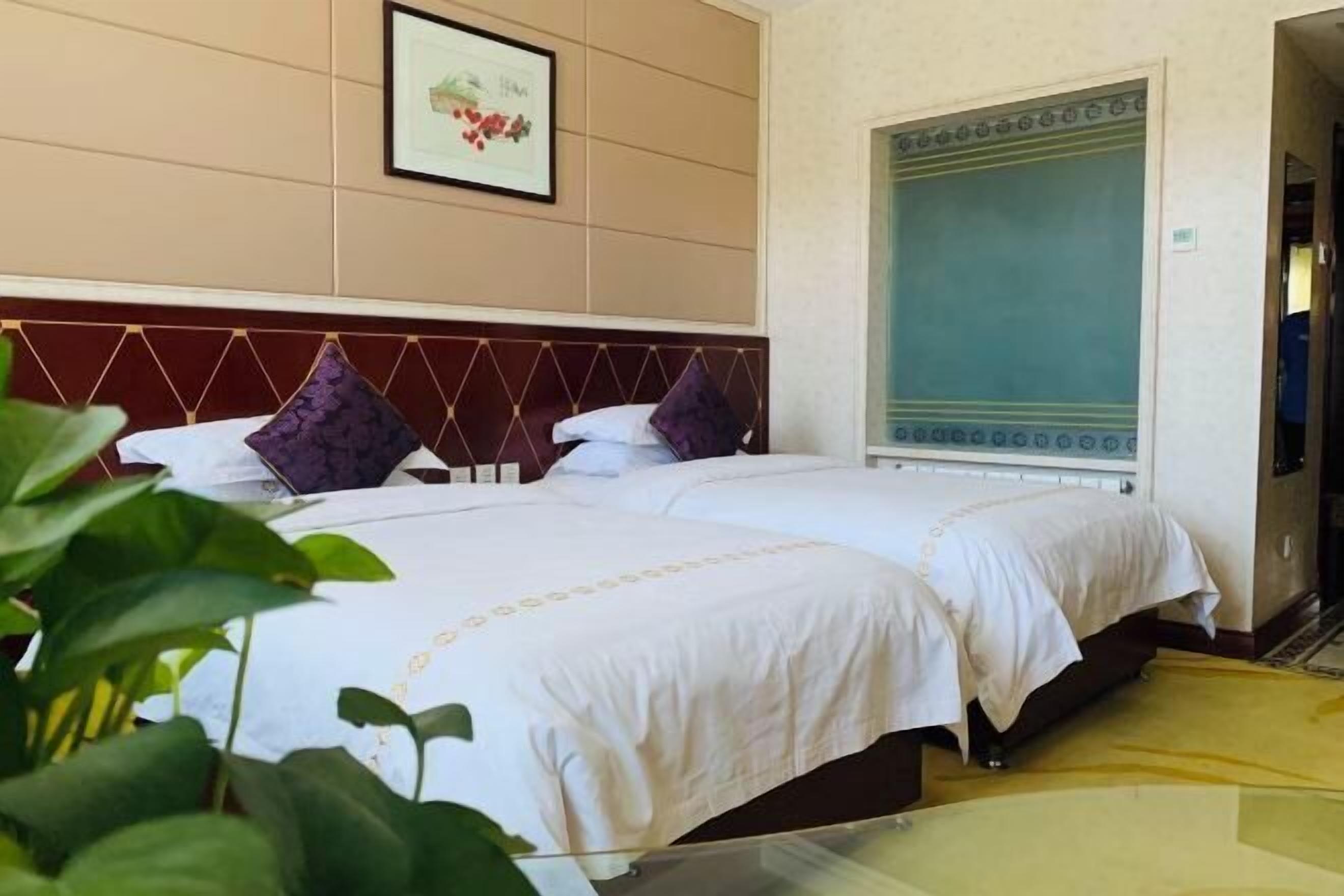 A hotel room with two beds, a large window with a view, a framed picture on the wall, and a plant in the foreground.