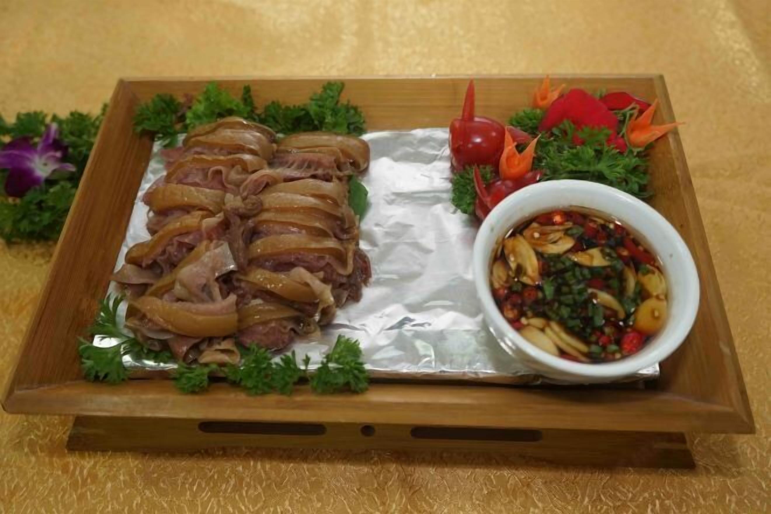 A wooden tray with sliced meat and a bowl of sauce, garnished with parsley and decorative vegetables.