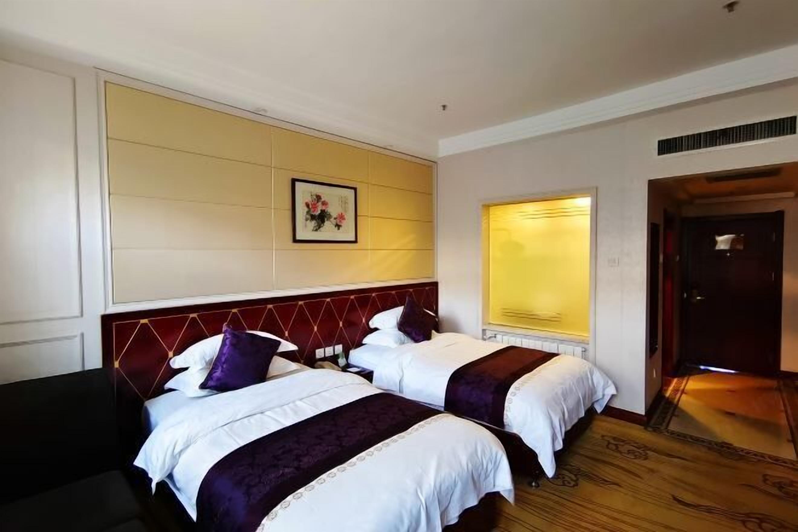 Ein Hotelzimmer mit zwei Betten, einem dunkelbraunen Sofa, einem großen Fenster mit Jalousien und einem gerahmten Kunstwerk an der Wand.