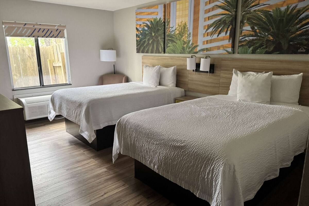 Habitación de hotel con dos camas, piso de madera, ventana con persianas, lámpara de pared y un cuadro de palmeras.