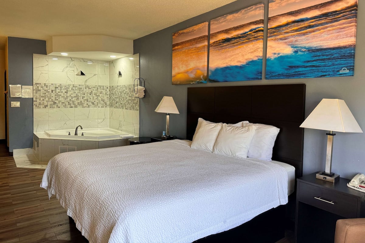Habitación de hotel con una cama grande, dos lámparas de noche, una mesita de noche, un baño con bañera, lavamanos y un espejo instalado en la pared.
