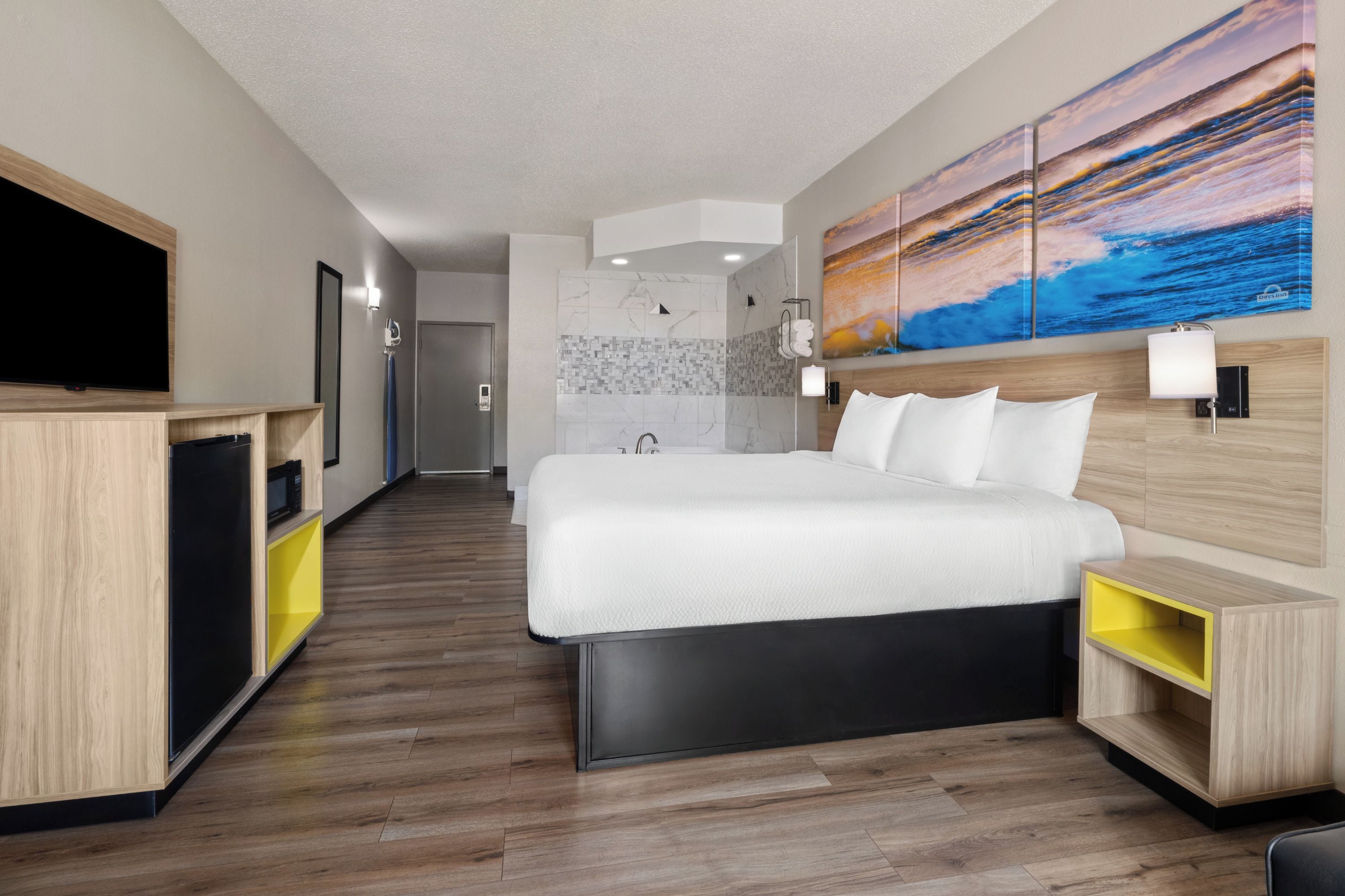 A modern hotel room with a large bed, a TV, a bathroom with a shower, and a painting on the wall.