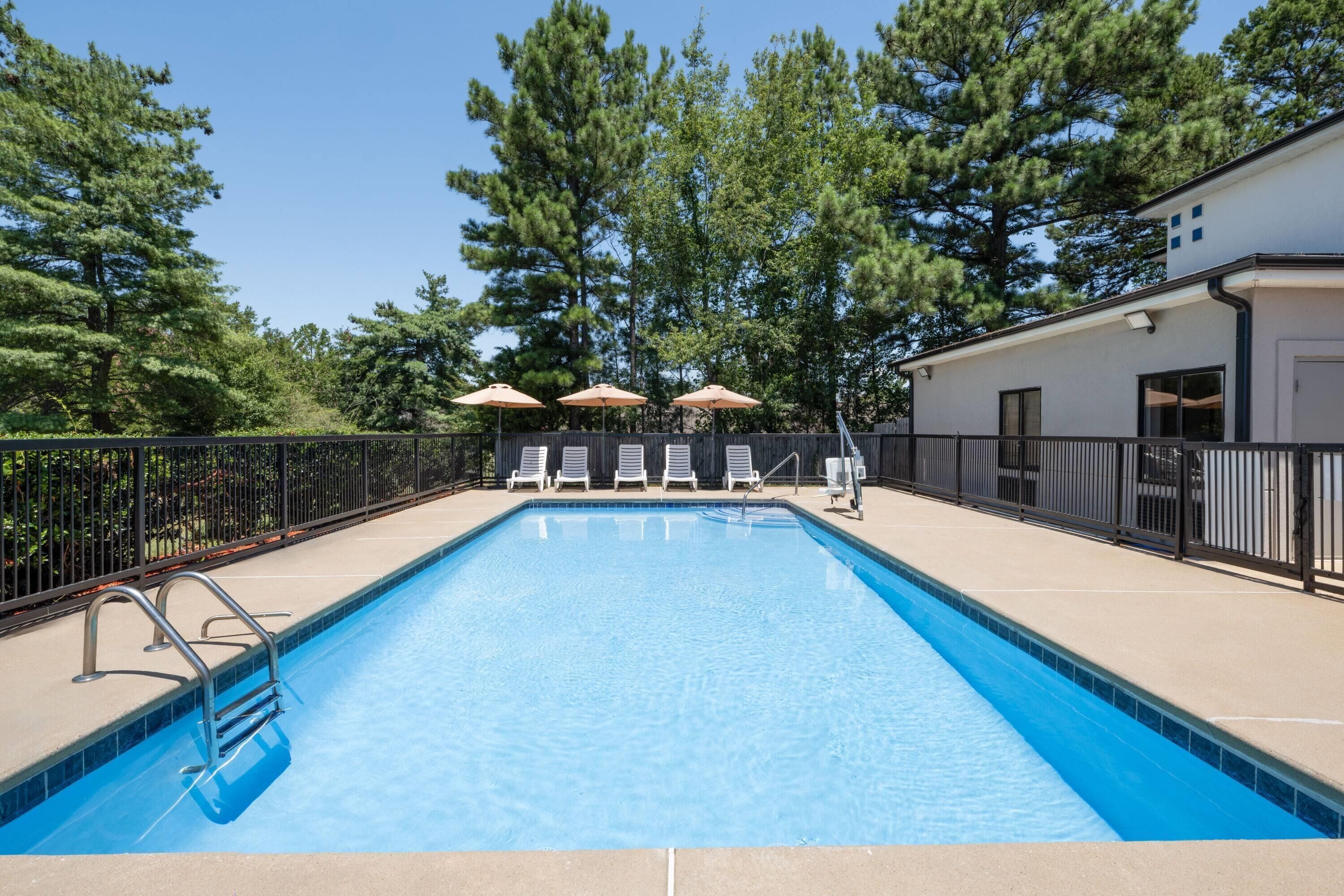 A rectangular swimming pool with clear blue water, surrounded by a black metal fence. There are three lounge chairs and two umbrellas under a wooden deck.