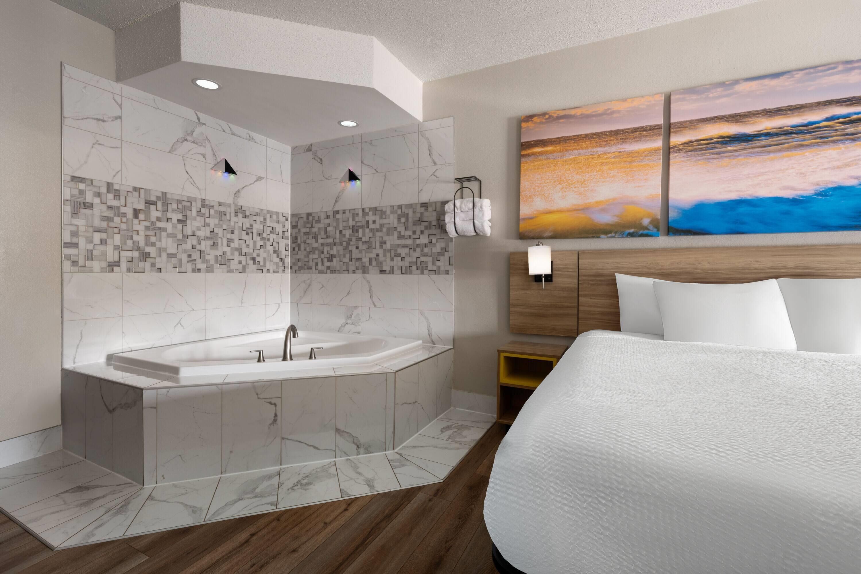 A modern hotel room with a large bed, a bathtub, and a wall-mounted painting.