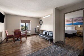 A hotel room with a wooden floor, a sofa, a desk with a chair, a bed, and a painting on the wall.