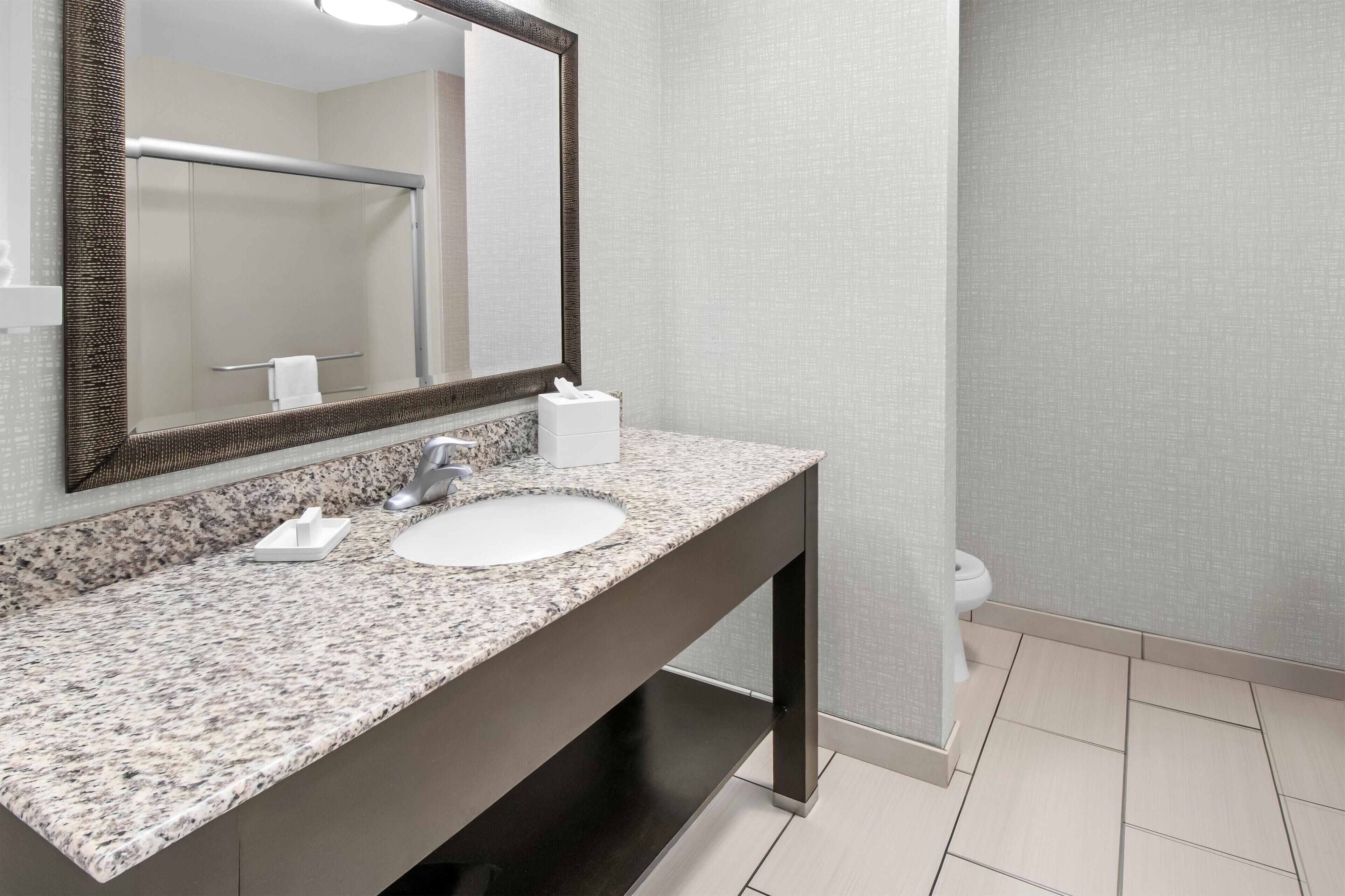 A modern bathroom with a granite countertop, a round sink, a large mirror, and a towel dispenser.