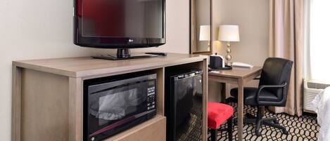 Habitación de hotel con televisión de pantalla plana, microondas, refrigerador pequeño, escritorio con silla, lámpara y cama con manta roja.