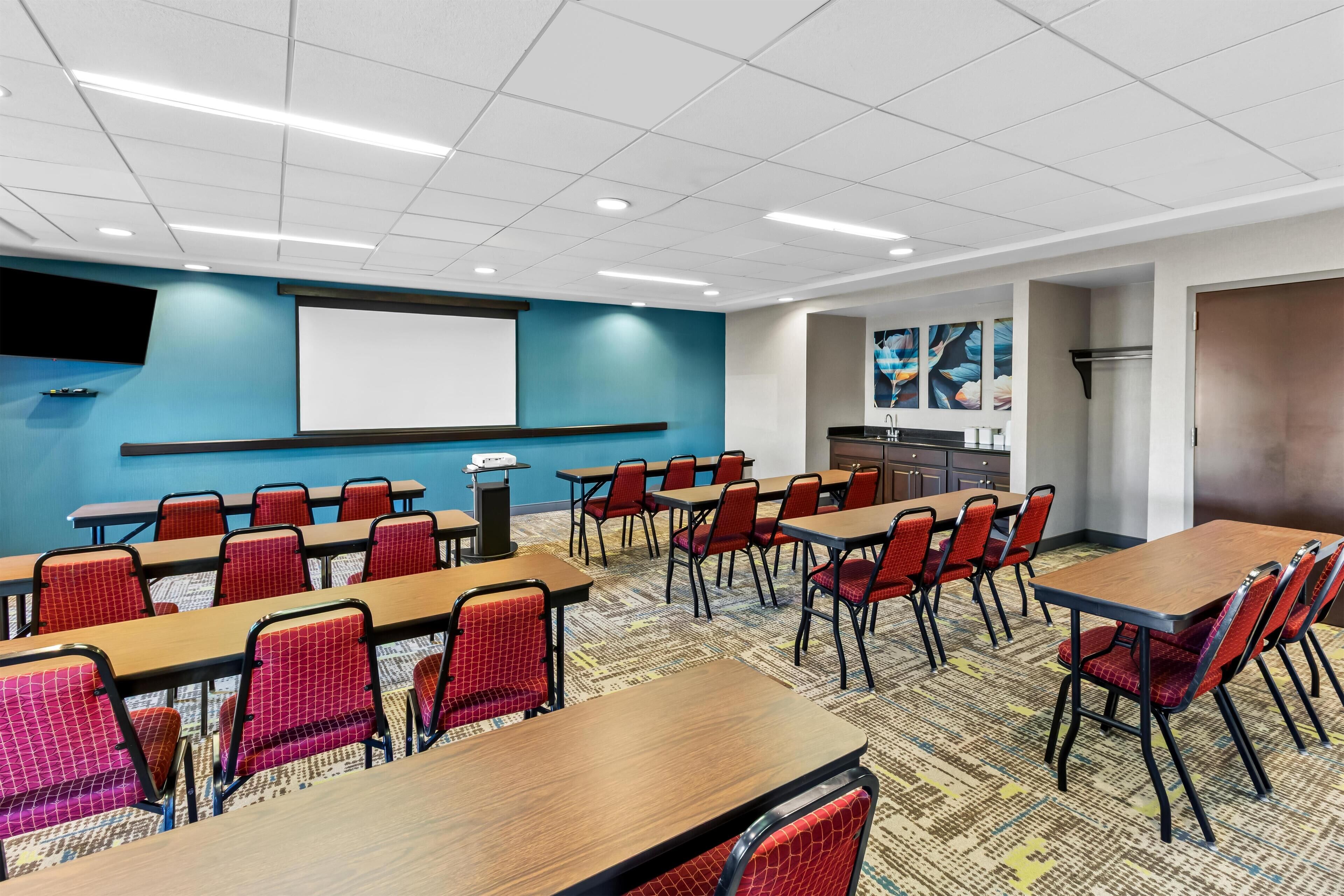 A conference room with multiple tables and chairs, a projector screen, and a carpeted floor.