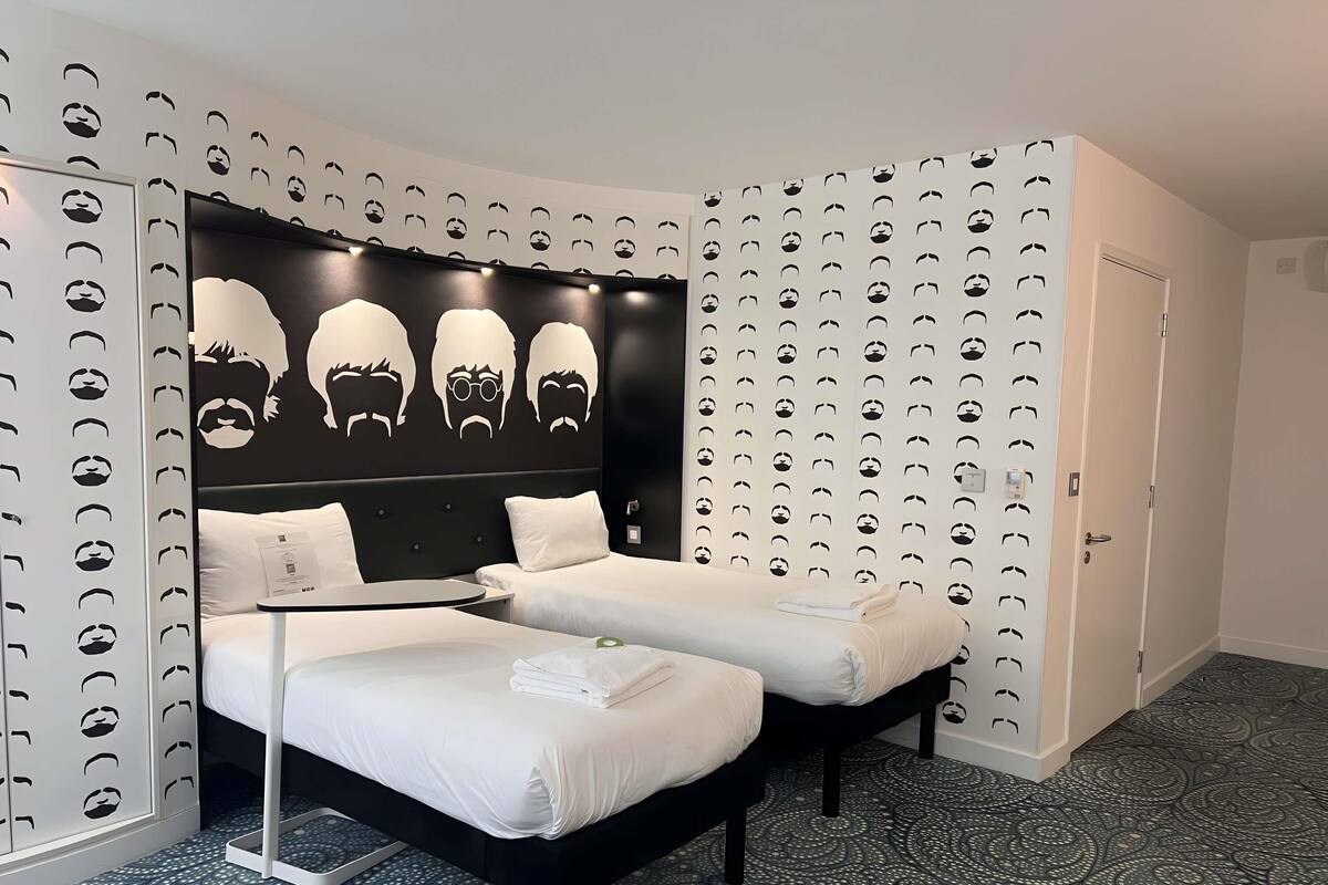 A hotel room with a bed, two bedside tables, a headboard with a unique design, patterned wallpaper, and a door.
