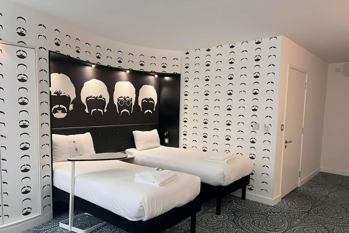 A hotel room with a bed, two bedside tables, a headboard with a unique design, patterned wallpaper, and a door.
