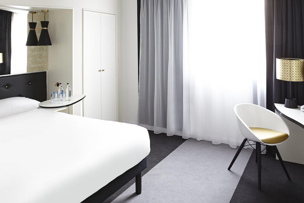 A hotel room with a bed, a desk, a chair, a mirror, and a window with curtains.