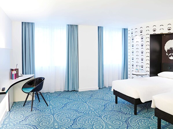 A hotel room with a bed, a desk with a chair, a large window with curtains, and a wall with a pattern of faces.