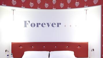 Hotellihuone, jossa on punainen pääty, valkoiset vuodevaatteet ja seinä, jossa on sana ”Forever” ja pieniä sydänkoristeita.
