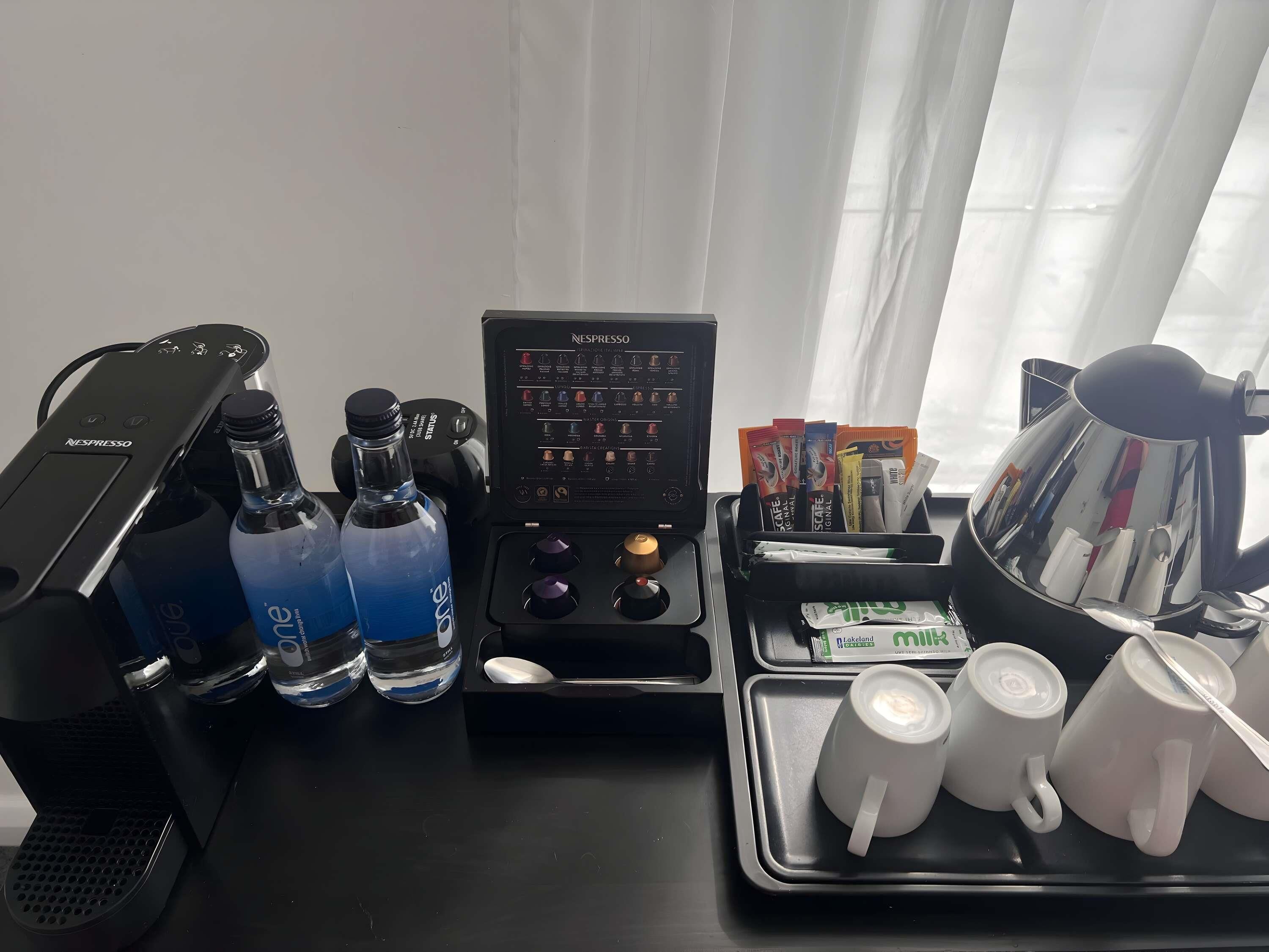 Una cafetera Nespresso, dos botellas de agua, un soporte para cápsulas de café y tres tazas blancas sobre una bandeja.