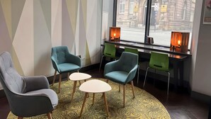 A modern interior with a seating area featuring two grey armchairs and three teal chairs around a small white table. A patterned rug covers the floor, and a large window offers a view of a historic building outside. - ibis Styles Liverpool Centre Dale Street (Liverpool)