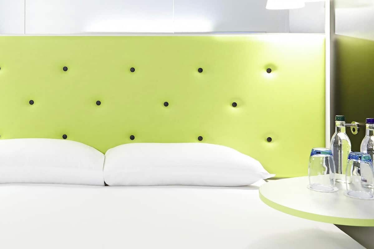 A bed with a green headboard, white pillows, and a bedside table with glasses and bottles.