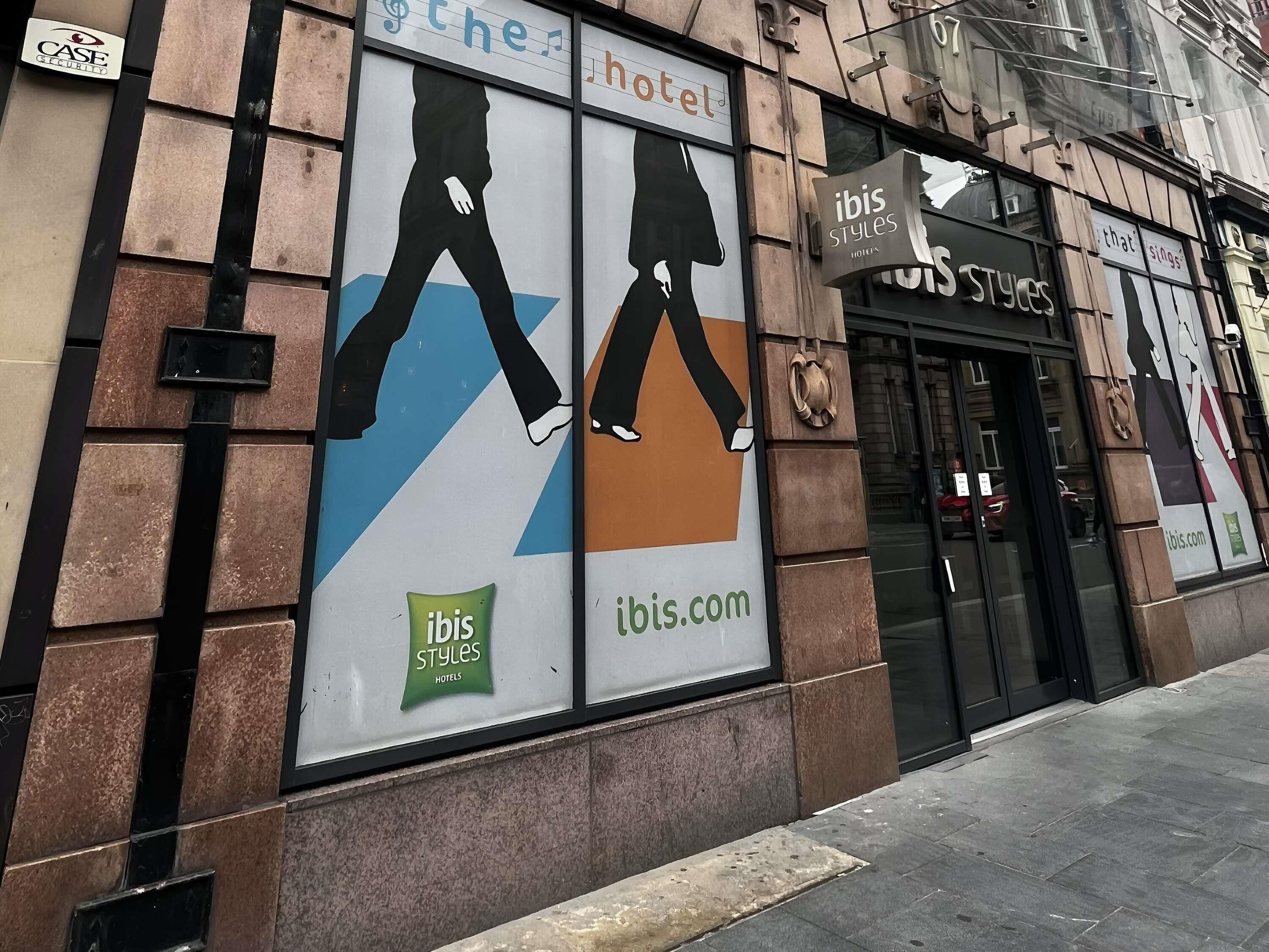A building facade with a large billboard advertising ibis Styles Hotels, featuring silhouettes of people walking.