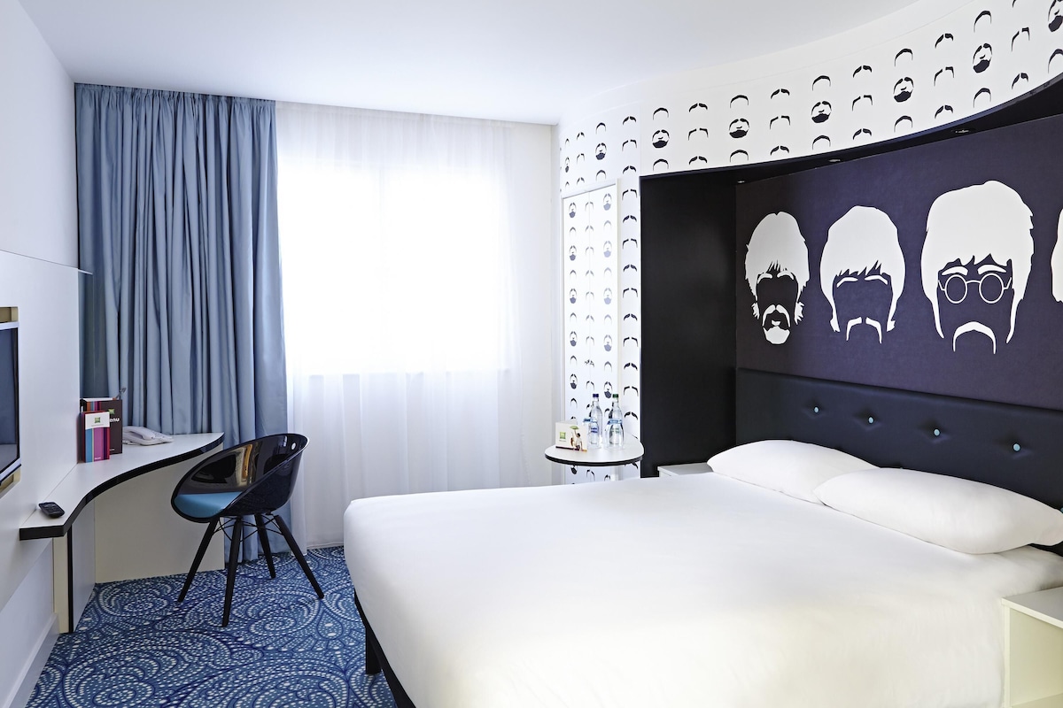A hotel room with a bed, a desk, a chair, and a wall with a pattern of faces.