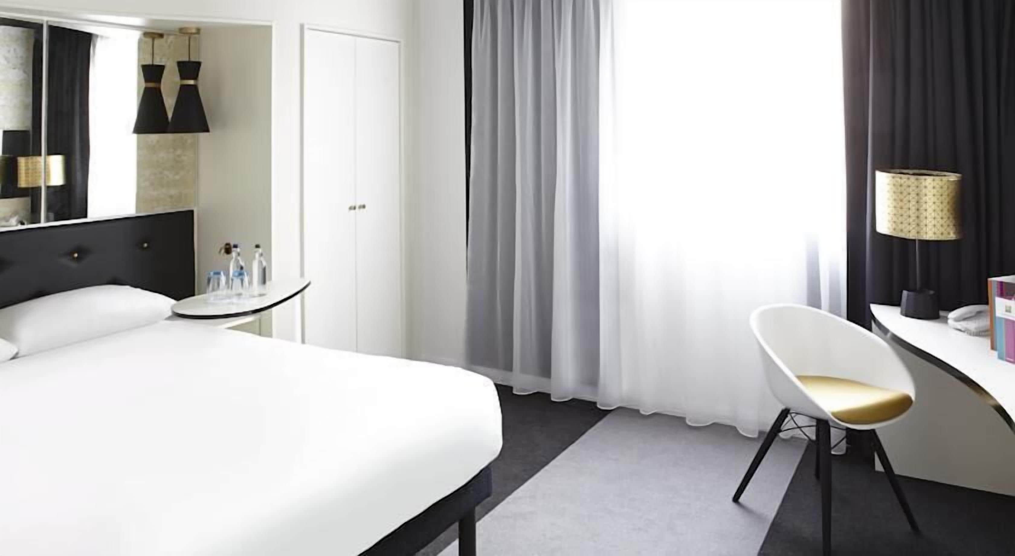 Quarto de hotel moderno com uma cama grande, uma escrivaninha com cadeira, um espelho e uma janela com cortinas.