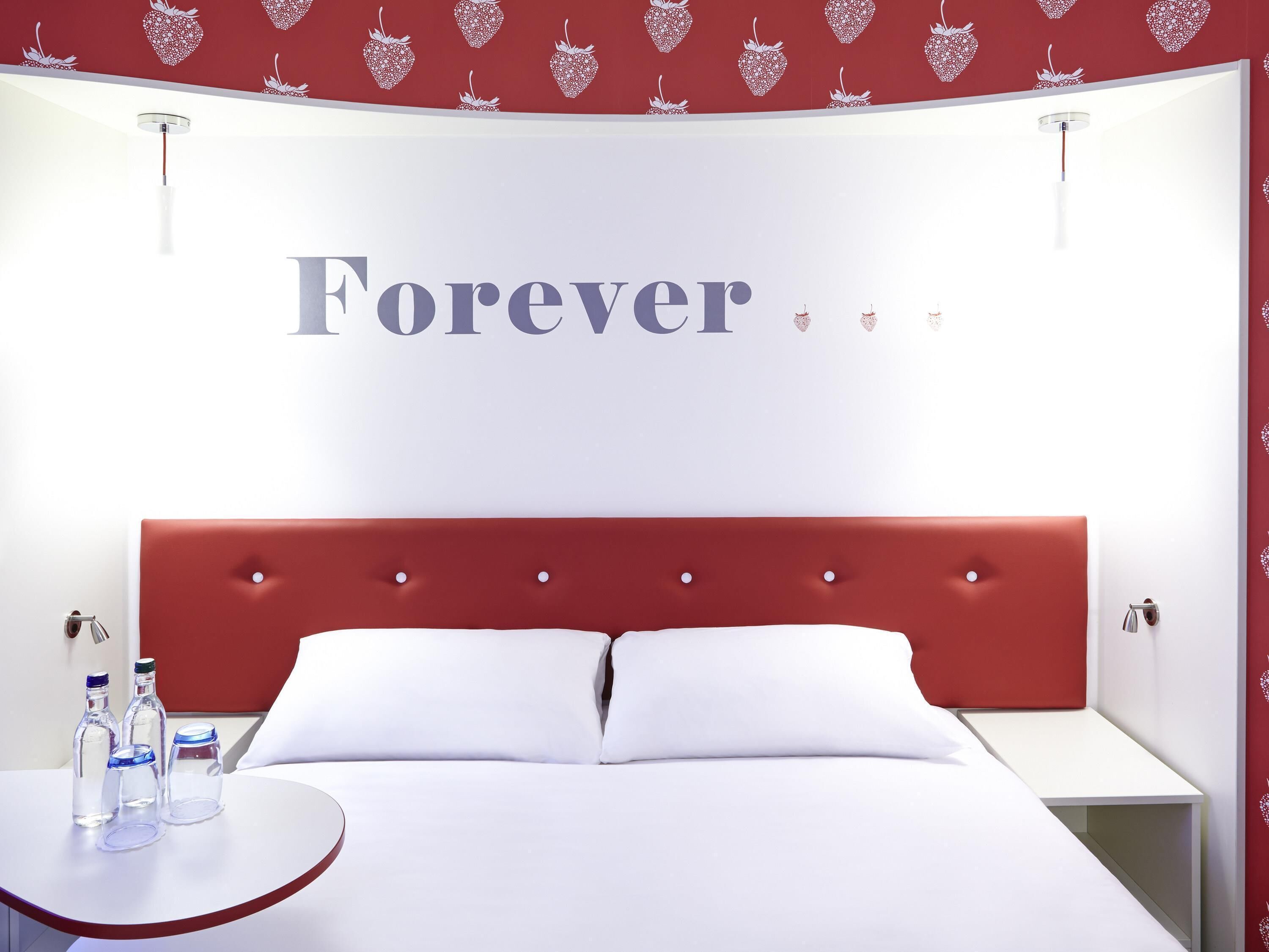 A hotel room with a red headboard, white bedding, and a wall with the word 'Forever' and heart patterns.