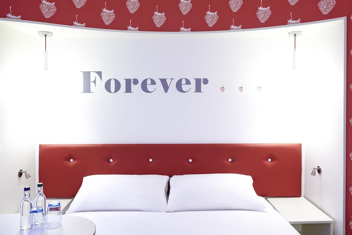 A hotel room with a red headboard, white bedding, and a wall with the word 'Forever' and heart patterns.