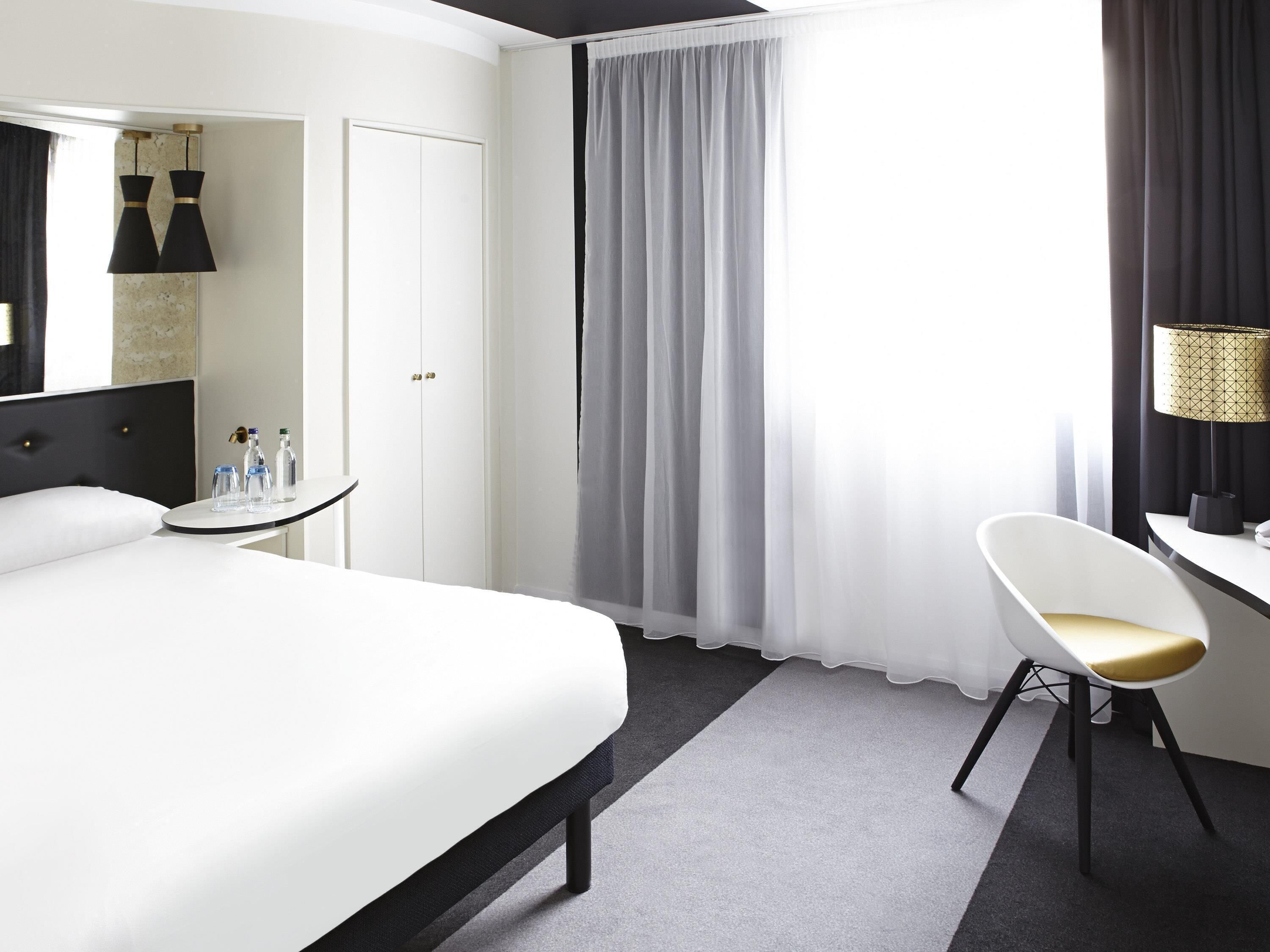 A hotel room with a bed, a desk, a chair, a lamp, and a window with curtains.