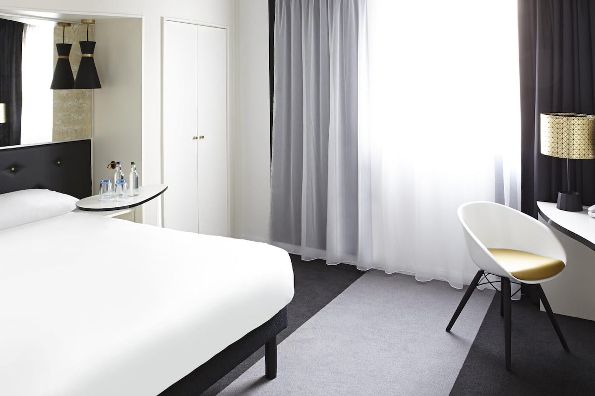 A hotel room with a bed, a desk, a chair, a lamp, and a window with curtains.