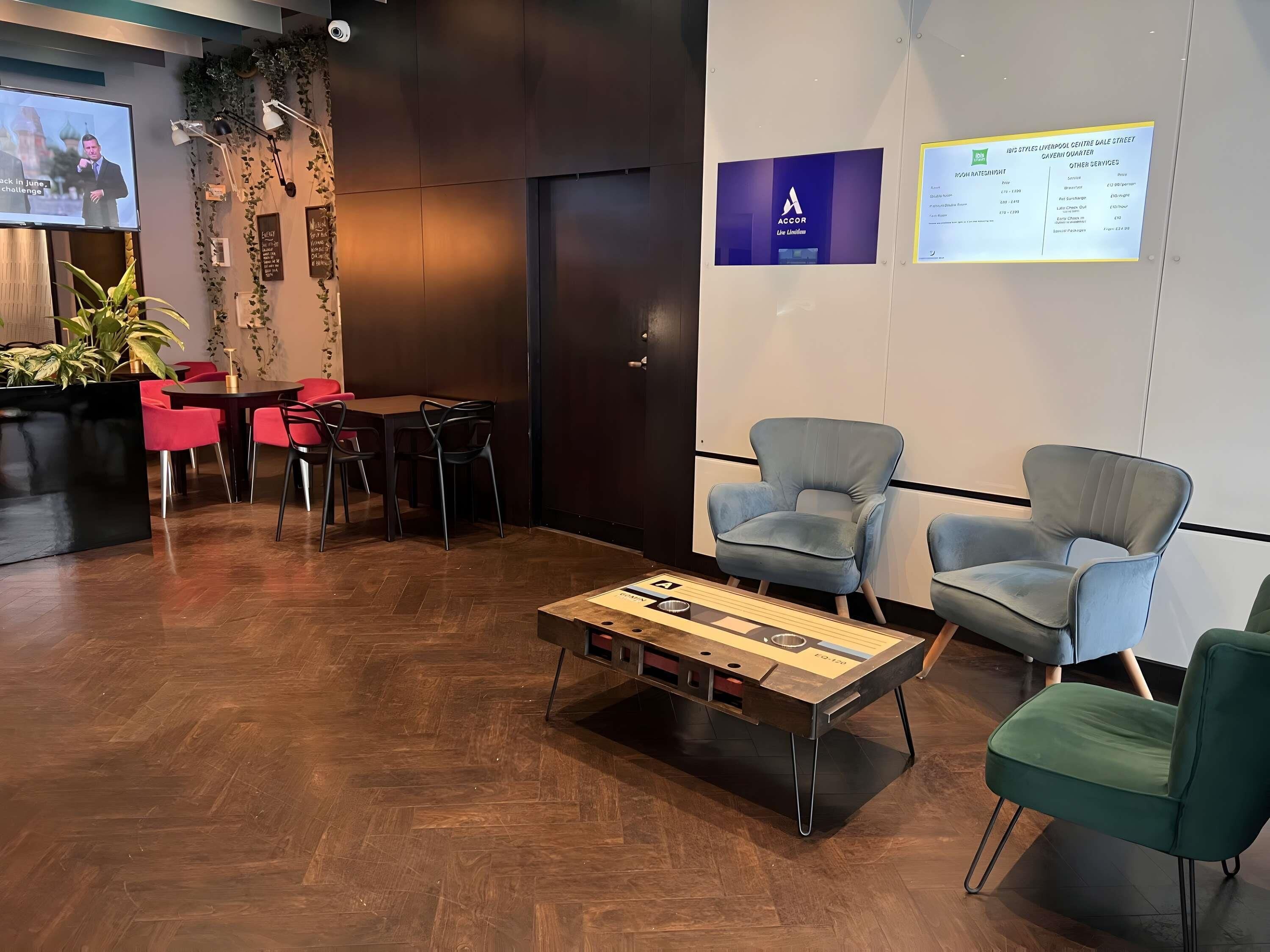 Un espace d’accueil avec une table basse en bois, des chaises rouges et noires, un écran de télévision et un mur orné d’affiches informatives.