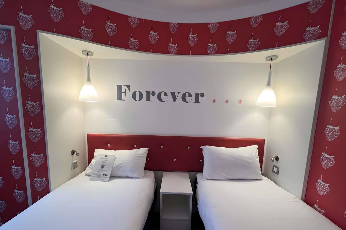 A hotel room with two beds, a red headboard, and a wall with the word 'Forever' and strawberry patterns.