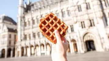 A hand holding a waffle in front of a historic building with arches and a tower.