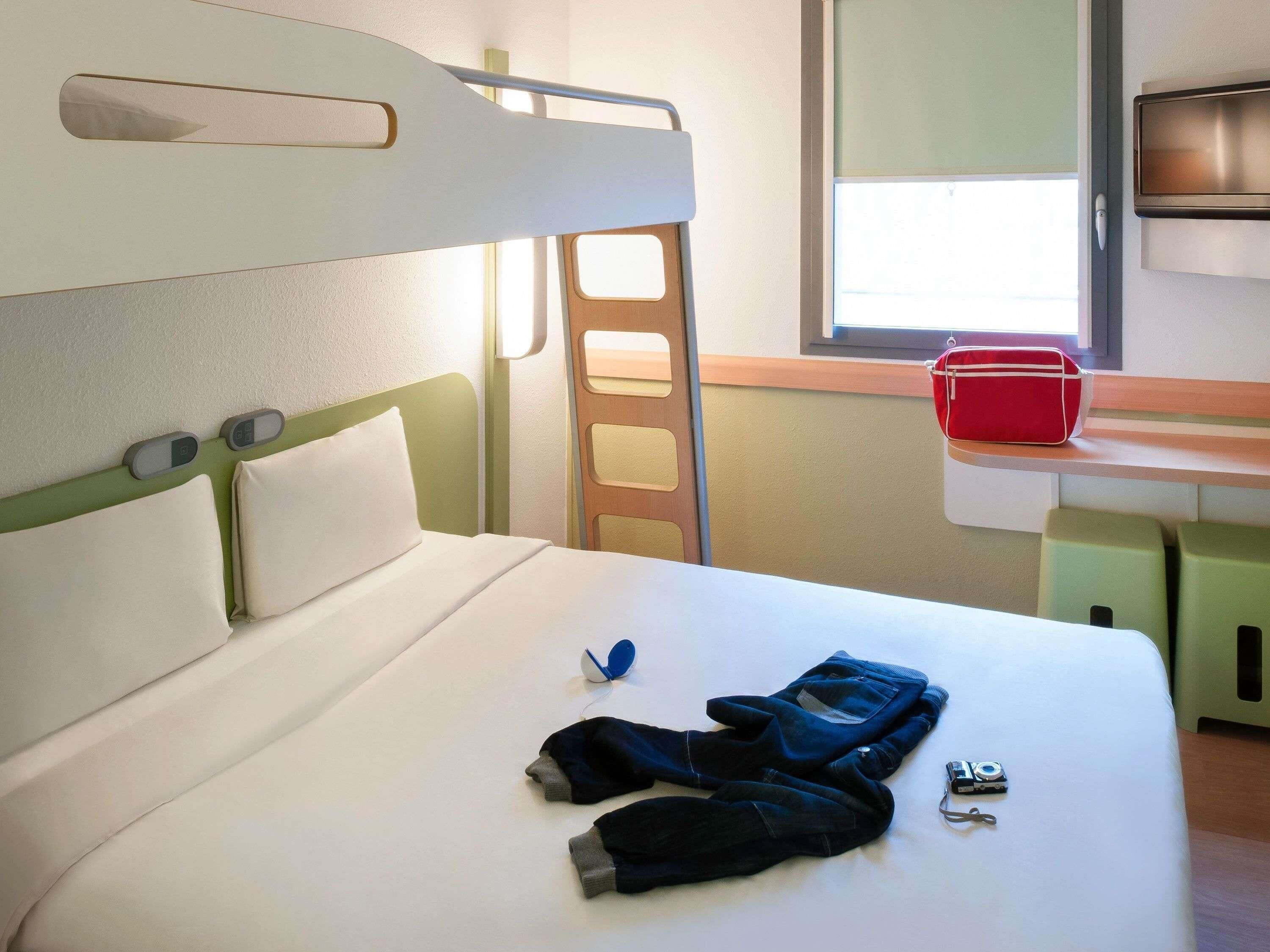 A bunk bed with a green headboard, a bedside table with a red bag, and a window with daylight.