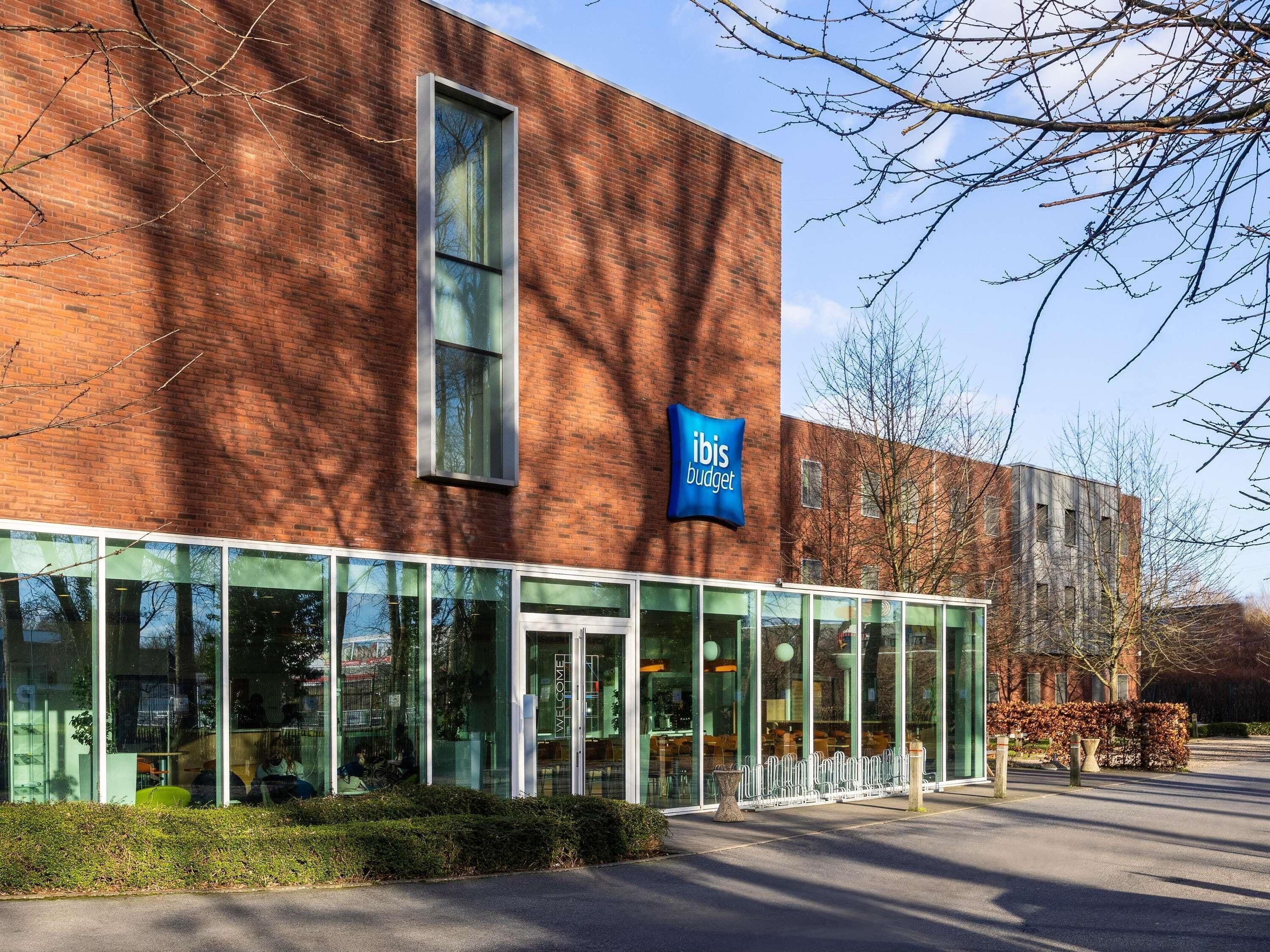 Een modern gebouw met een glazen gevel, een bord waarop 'ibis budget' staat, en een heldere lucht.