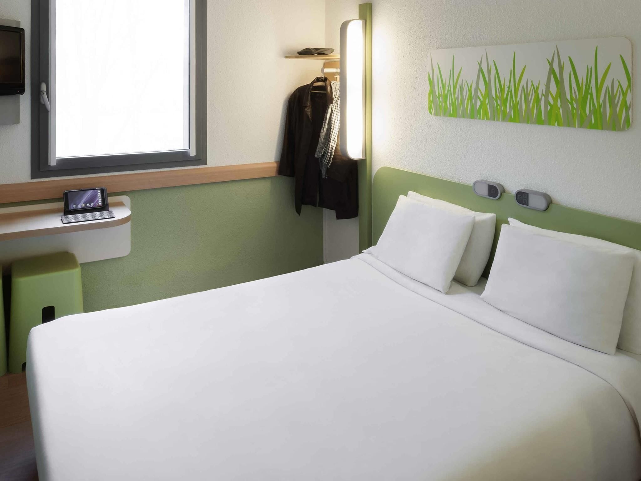 A hotel room with a bed, a small desk with a laptop, a coat rack, and a wall-mounted artwork.