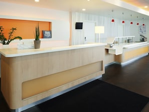 A modern hotel reception area with a wooden reception desk, a potted plant, a sign for ibis budget hotels, a flat-screen TV, and a counter with a lamp.