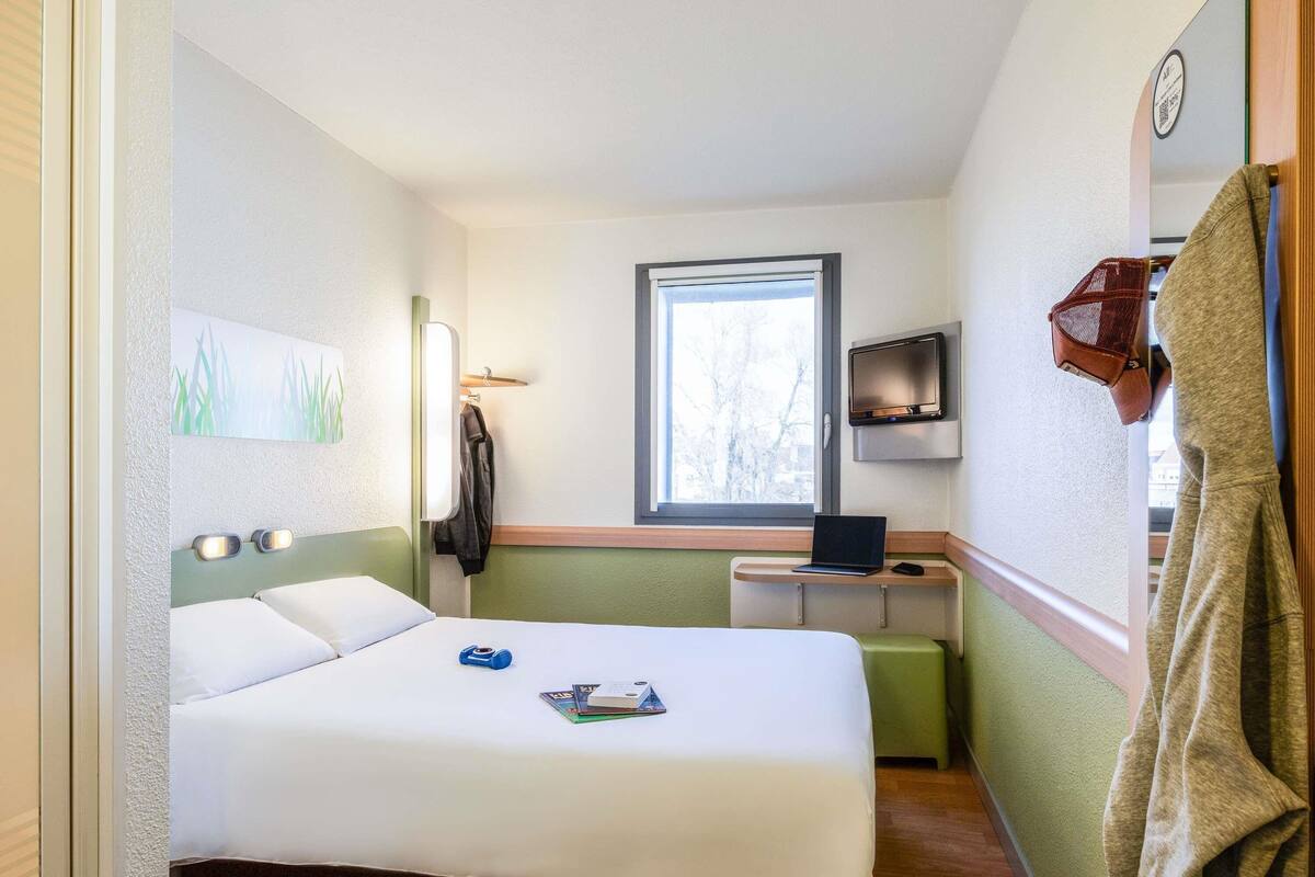 A compact hotel room with a bed, a desk with a laptop, a TV, and a window.
