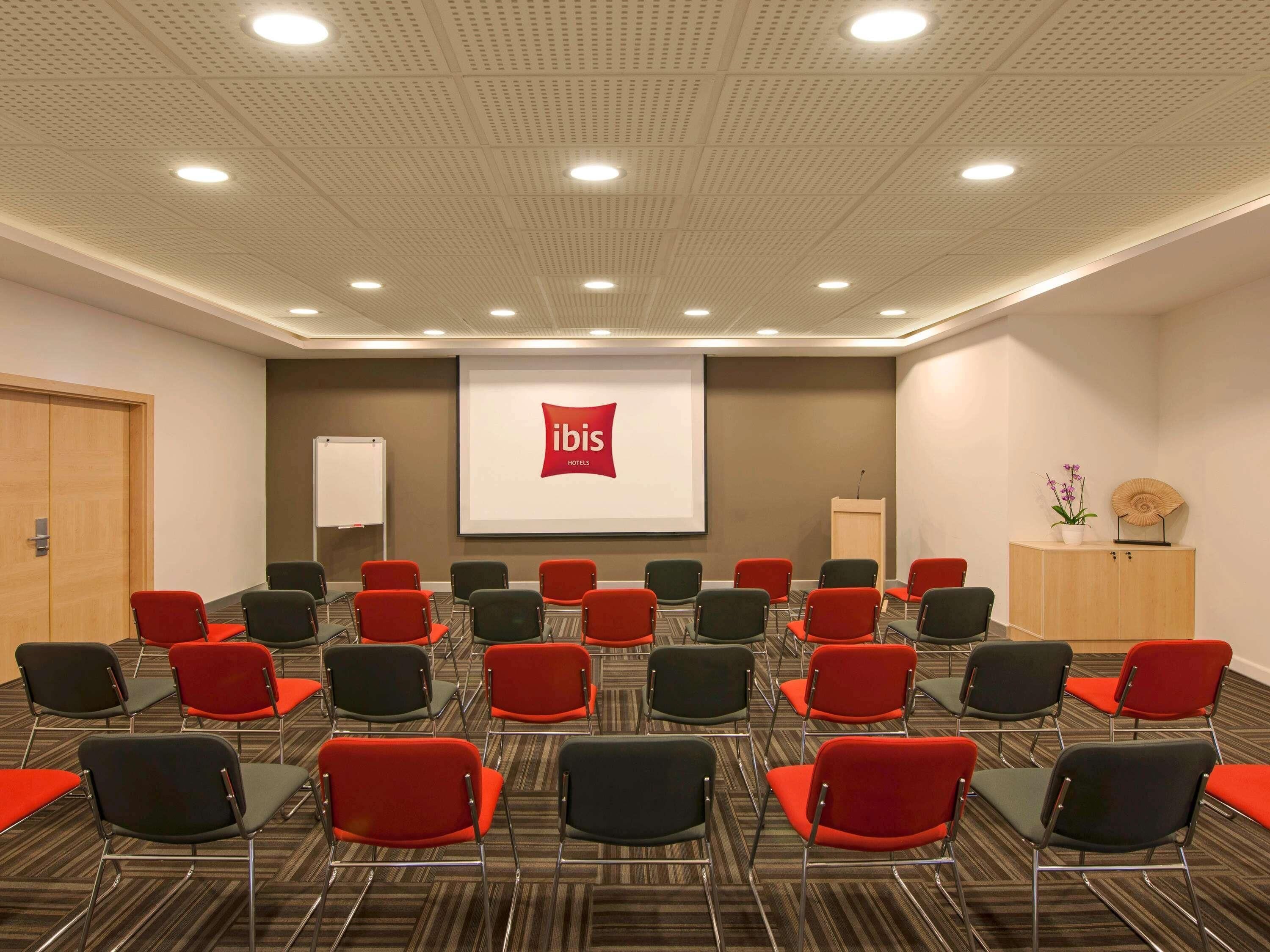 A conference room with rows of chairs, a projector screen displaying the ibis logo, a whiteboard, and a podium.