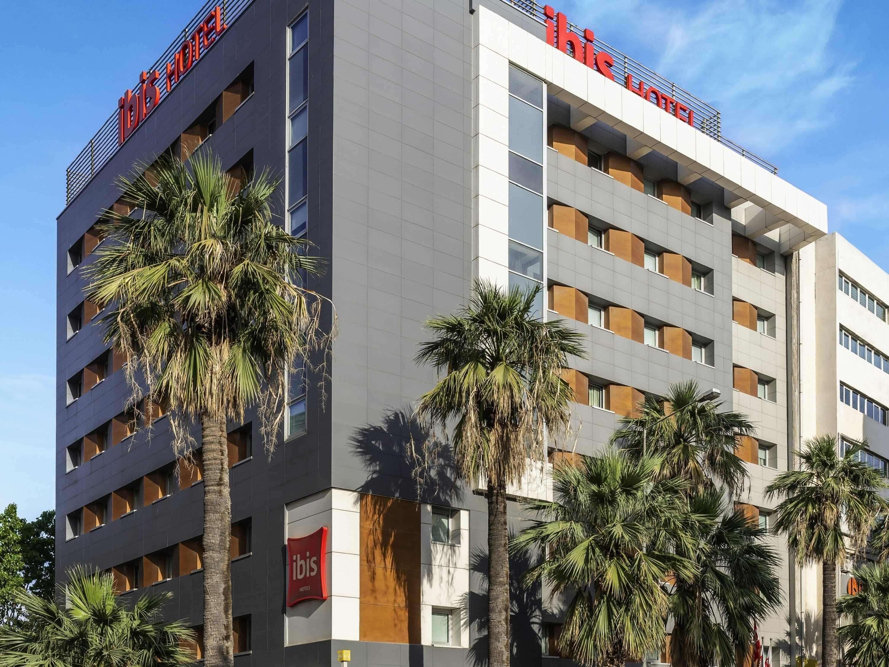 A modern hotel building with multiple floors, large windows, and palm trees in front.