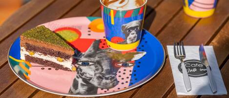 Una porción de pastel en un plato colorido, una taza de café, un tenedor y una servilleta con un logotipo sobre una mesa de madera.