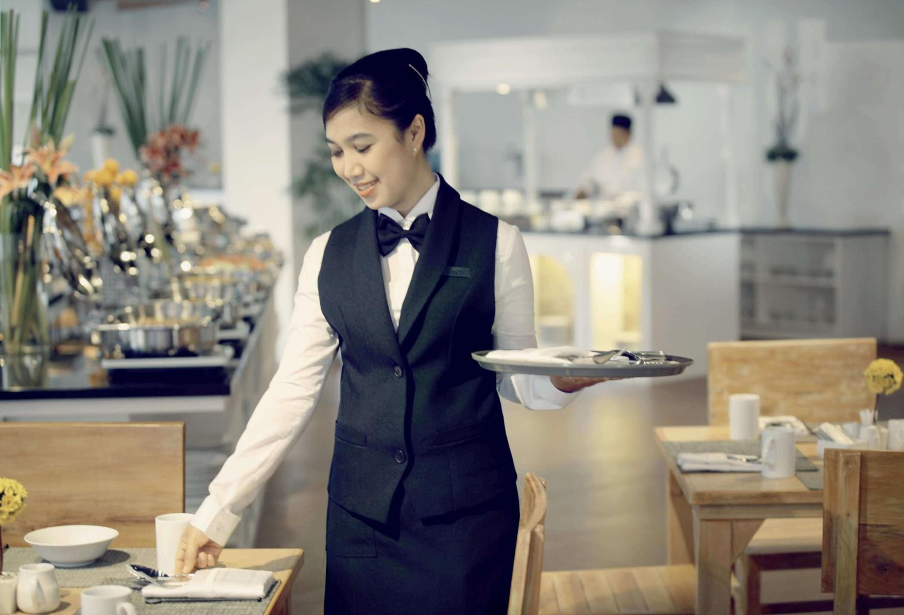 Un serveur, vêtu d’un uniforme élégant, porte un plateau chargé d’assiettes et d’une tasse.
