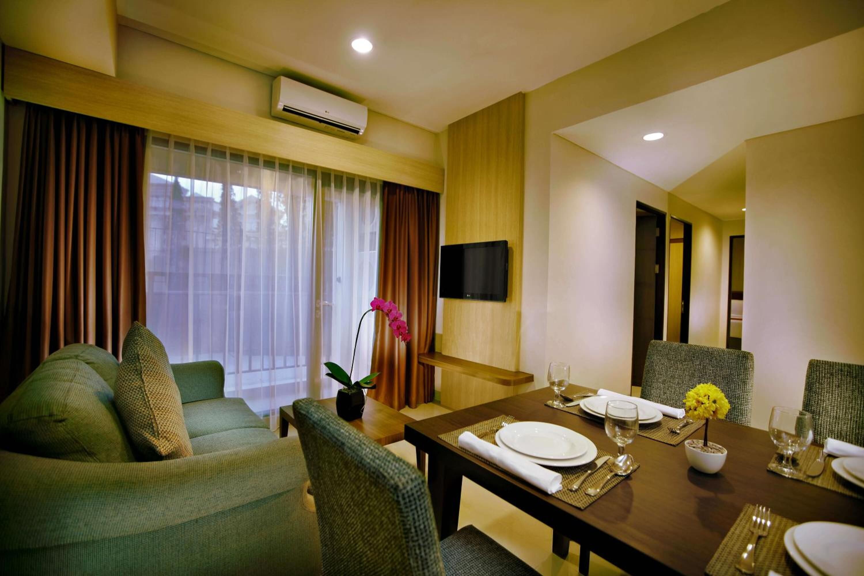 A hotel room with a dining area, a sofa, a TV mounted on the wall, and a potted plant.