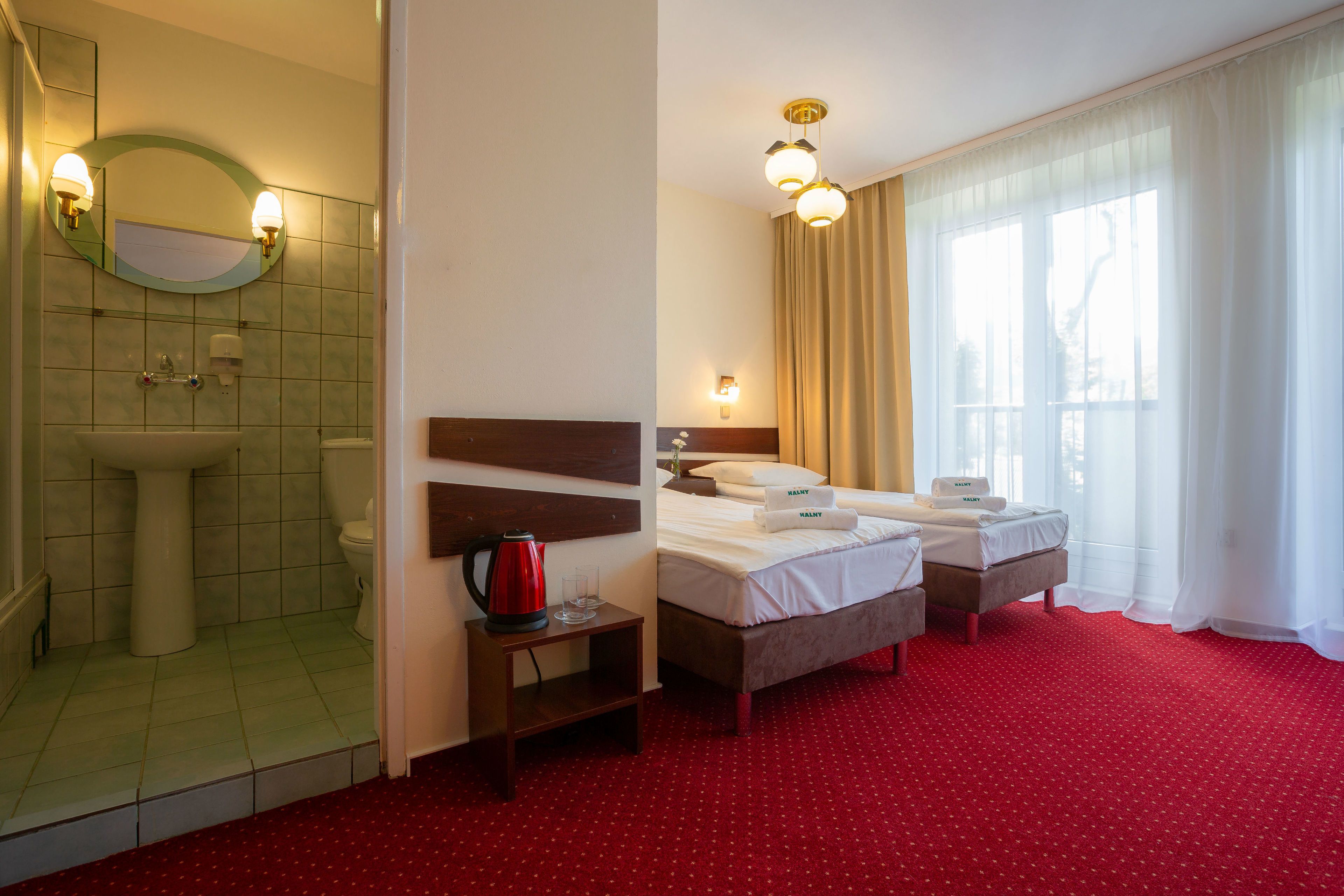 A hotel room with a red carpet, a bed with a wooden headboard, a bedside table with a red kettle, a mirror with wall sconces, and a window with sheer curtains.