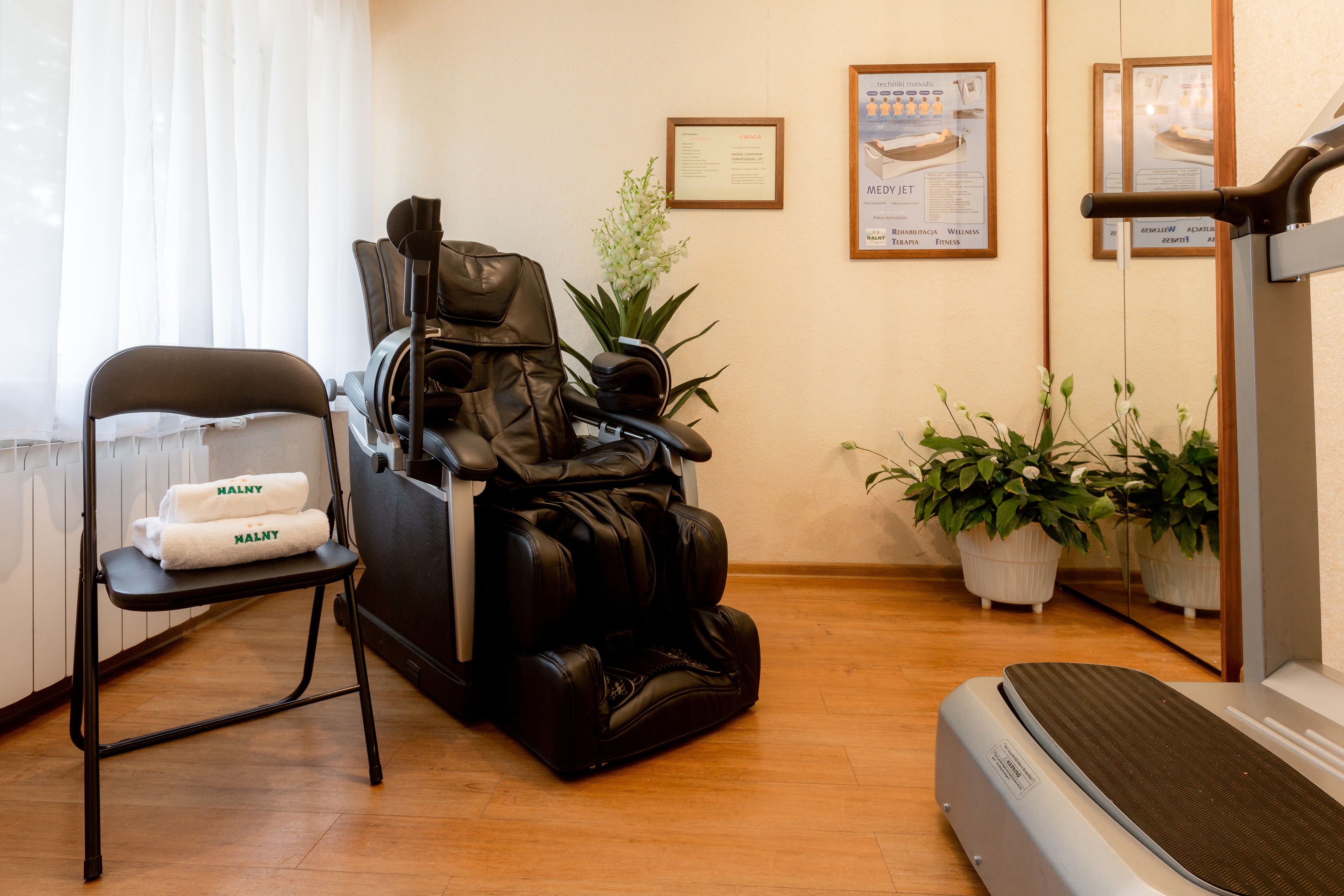 A massage therapy room with a massage chair, a stepper, a small table with towels, and potted plants.