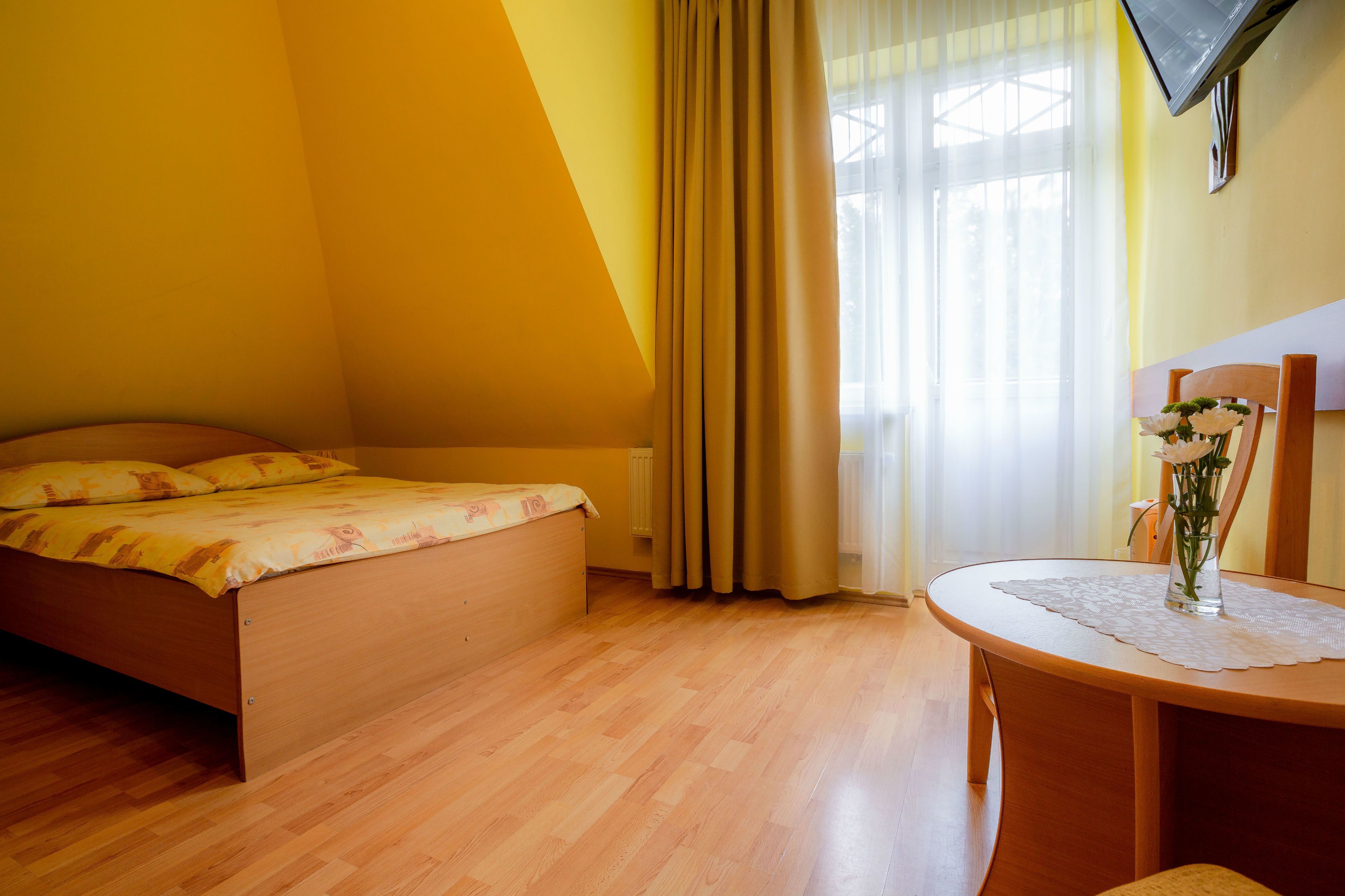 Un dormitorio con una cama, una mesa redonda de madera con un florero y una ventana con cortinas.