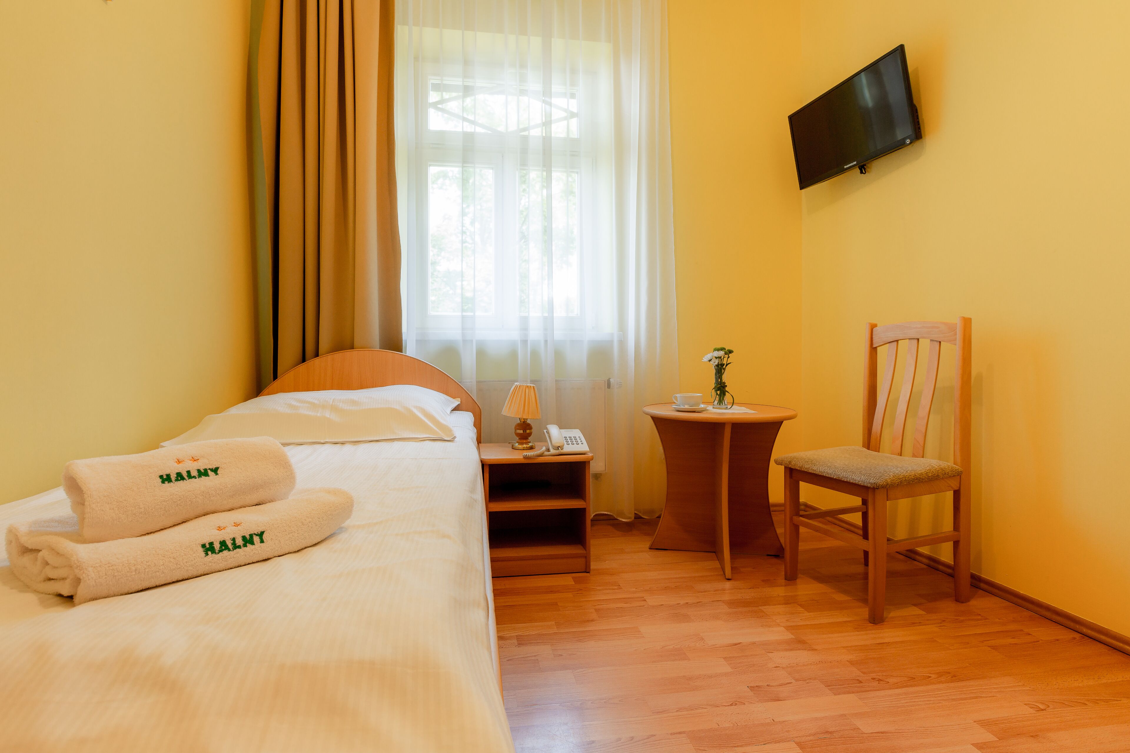 A hotel room with a bed, a chair, a small table, a nightstand, a TV, and a window with curtains.