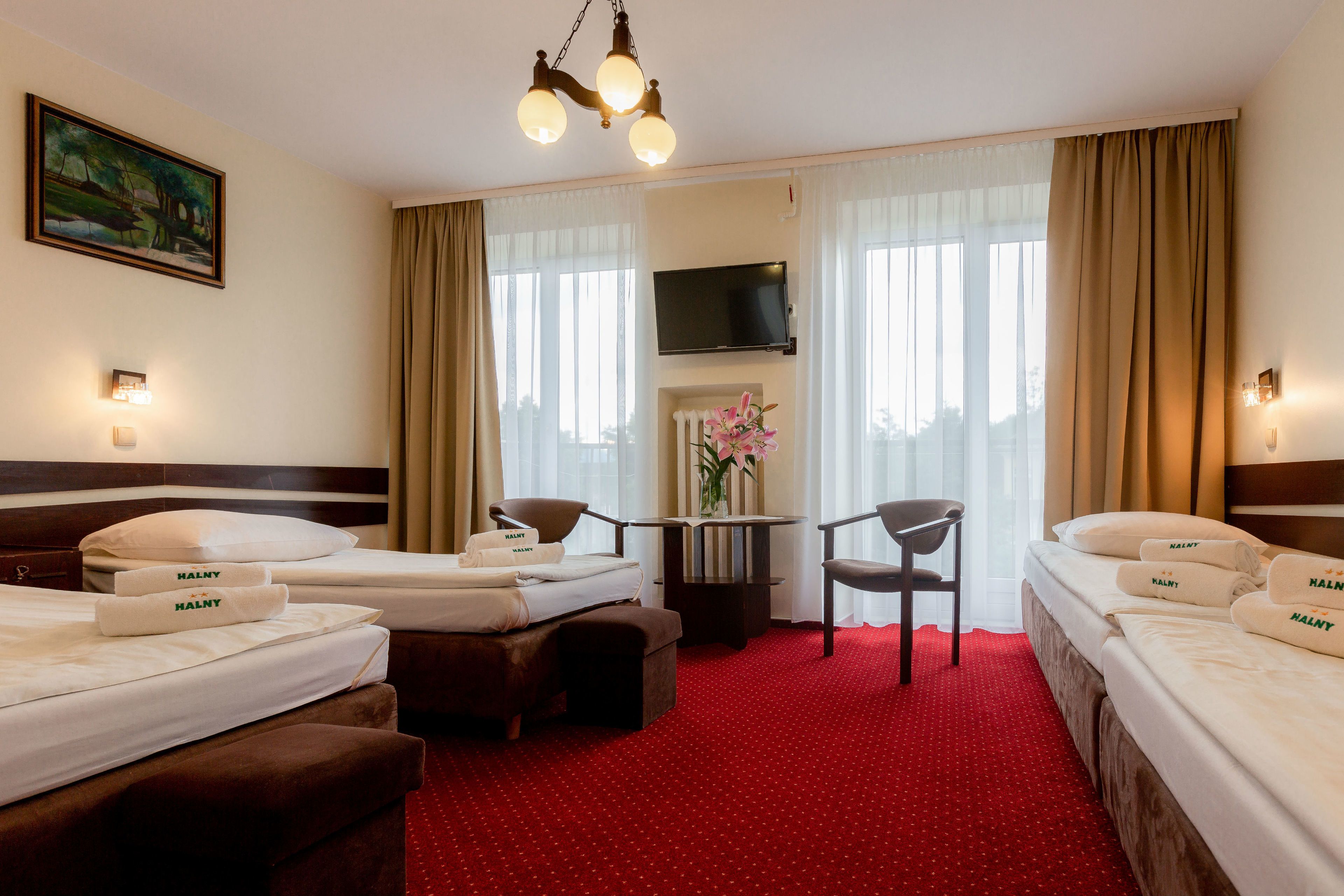 A hotel room with two beds, a TV, a table with chairs, and a red carpet.